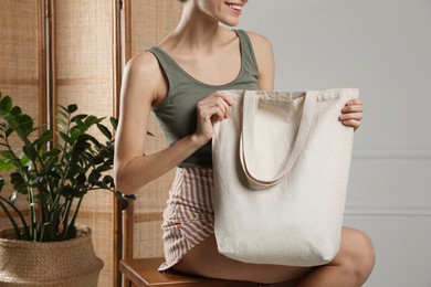 Photo of Happy young woman with blank eco friendly bag sitting on wooden stool indoors, closeup