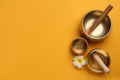 Photo of Golden singing bowls, mallets and flower on orange background, flat lay. Space for text