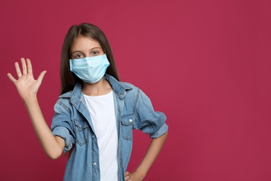 Photo of Little girl in protective mask showing hello gesture on crimson background, space for text. Keeping social distance during coronavirus pandemic