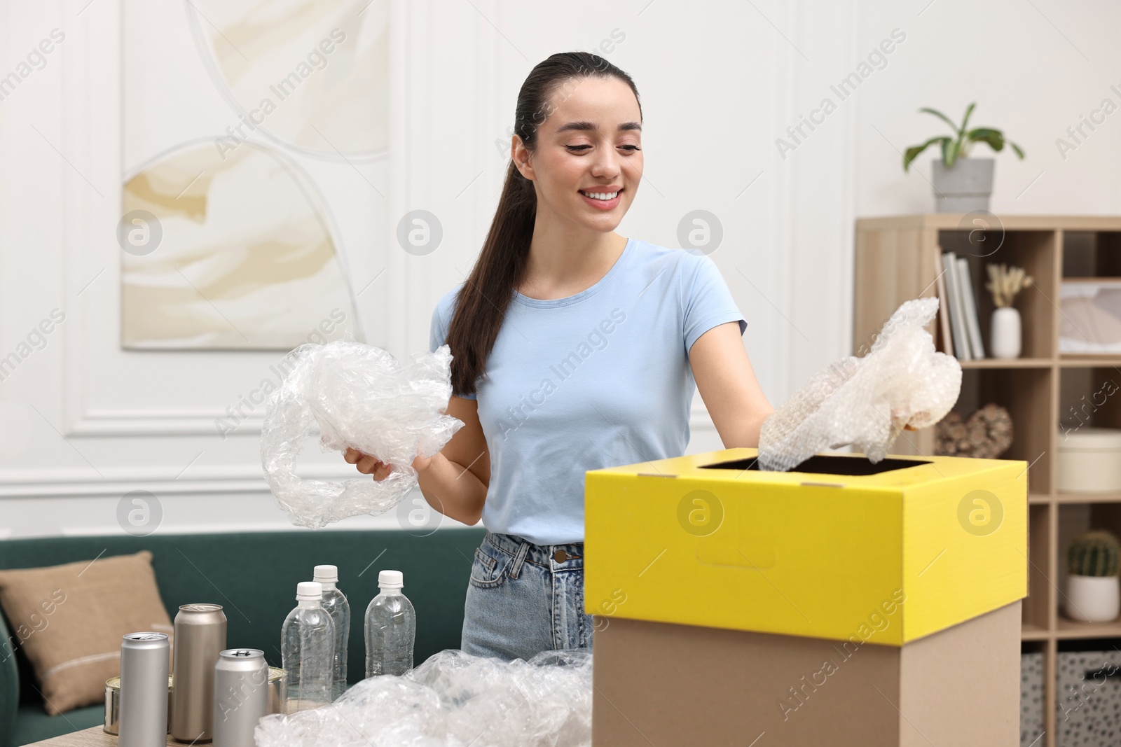 Photo of Garbage sorting. Smiling woman throwing plastic package into cardboard box in room