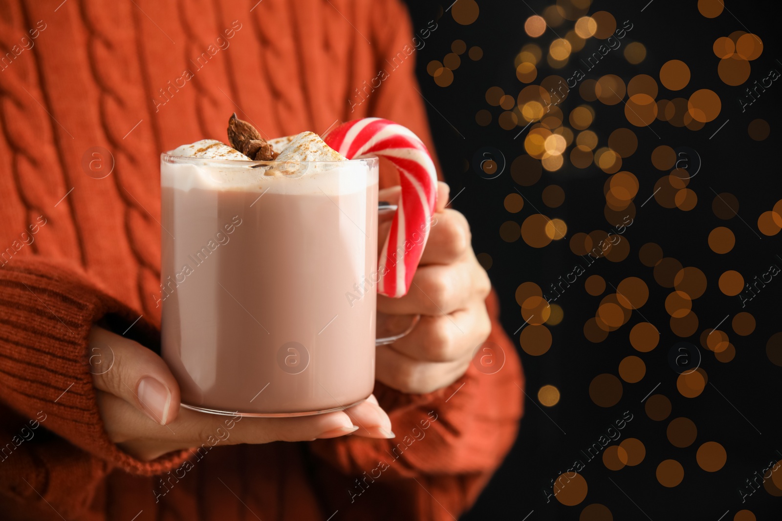 Photo of Woman holding glass cup of tasty cocoa with marshmallows and Christmas candy cane against blurred festive lights, closeup. Space for text