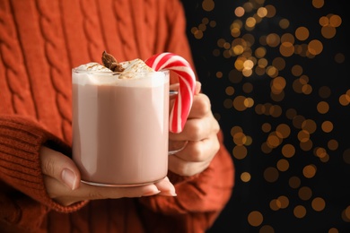 Photo of Woman holding glass cup of tasty cocoa with marshmallows and Christmas candy cane against blurred festive lights, closeup. Space for text