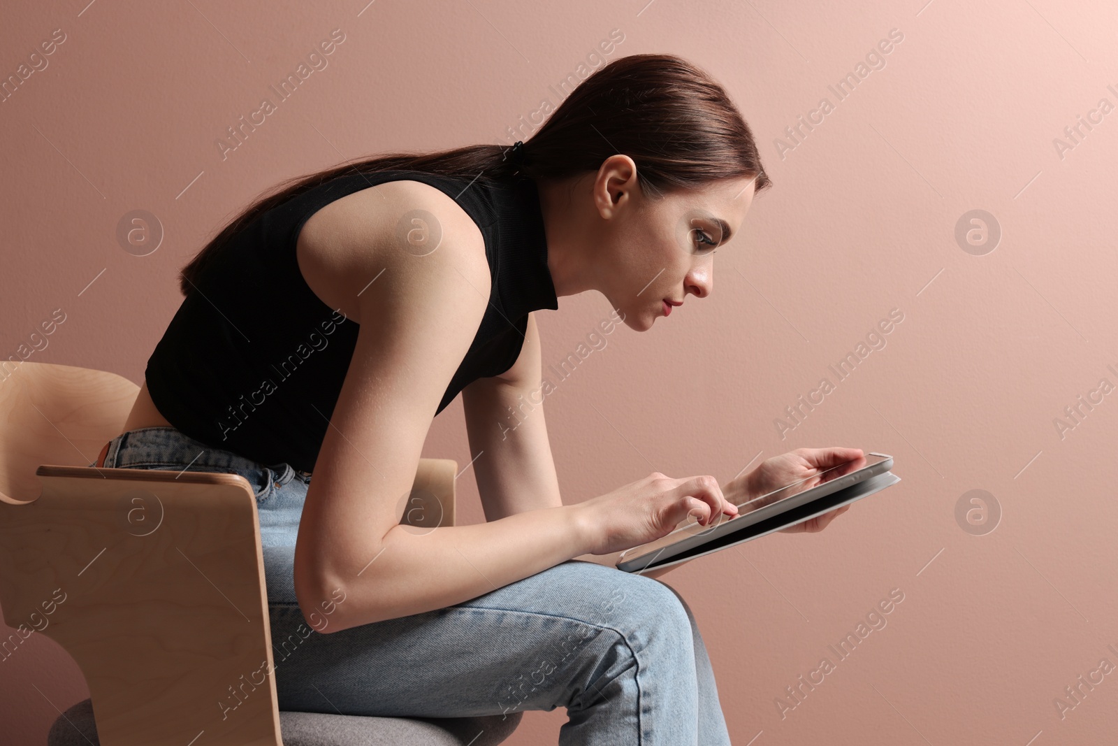 Photo of Young woman with bad posture using tablet while sitting on chair against pale pink background