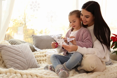 Photo of Mother and daughter making paper snowflake near window at home