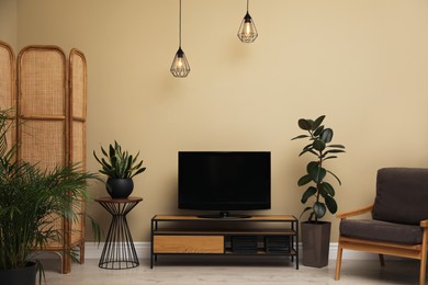 Photo of Modern TV on cabinet, armchair and green plants near beige wall in room. Interior design