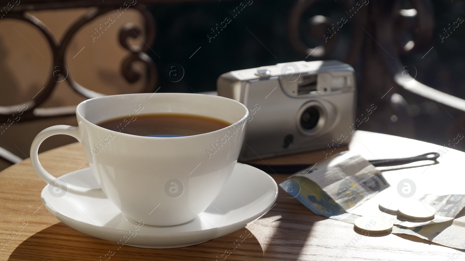 Photo of Cup of aromatic coffee, banknotes and camera on wooden table outdoors
