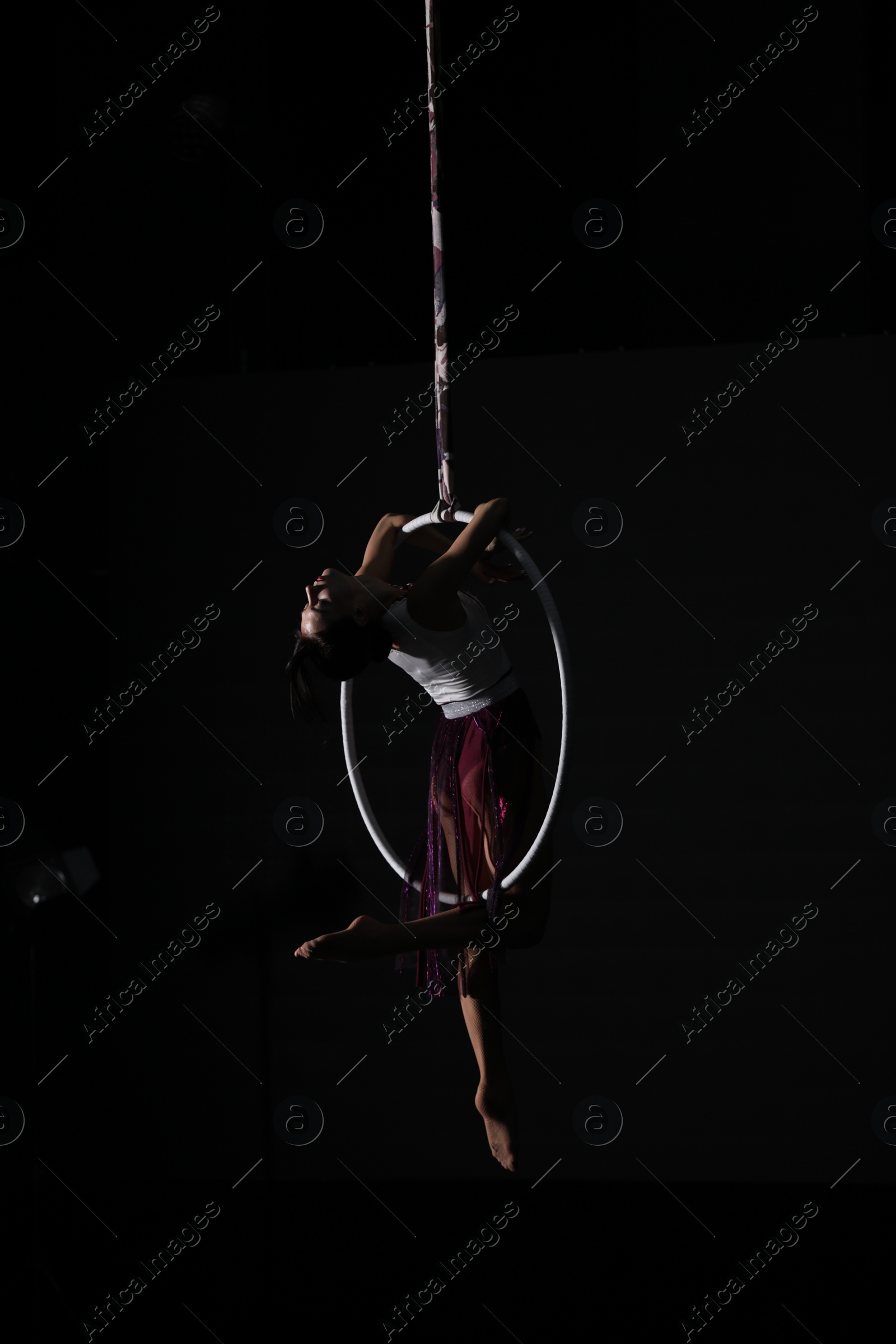 Photo of Young woman performing acrobatic element on aerial ring against dark background