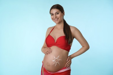 Young pregnant woman with sun protection cream on belly against light blue background