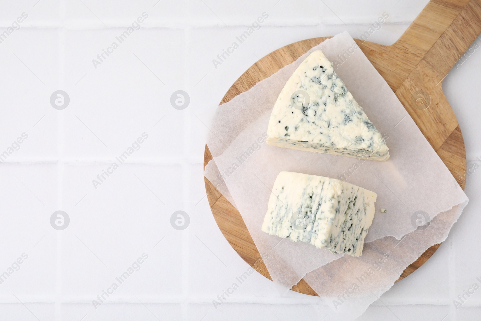 Photo of Wooden cutting board with blue cheese on white tiled table, top view. Space for text