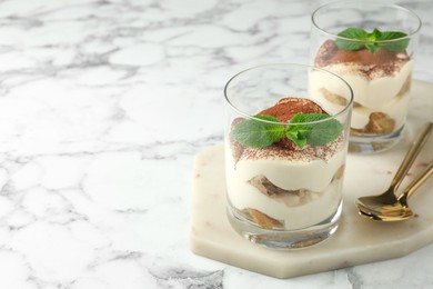 Photo of Delicious tiramisu in glasses, mint leaves and spoons on white marble table, space for text