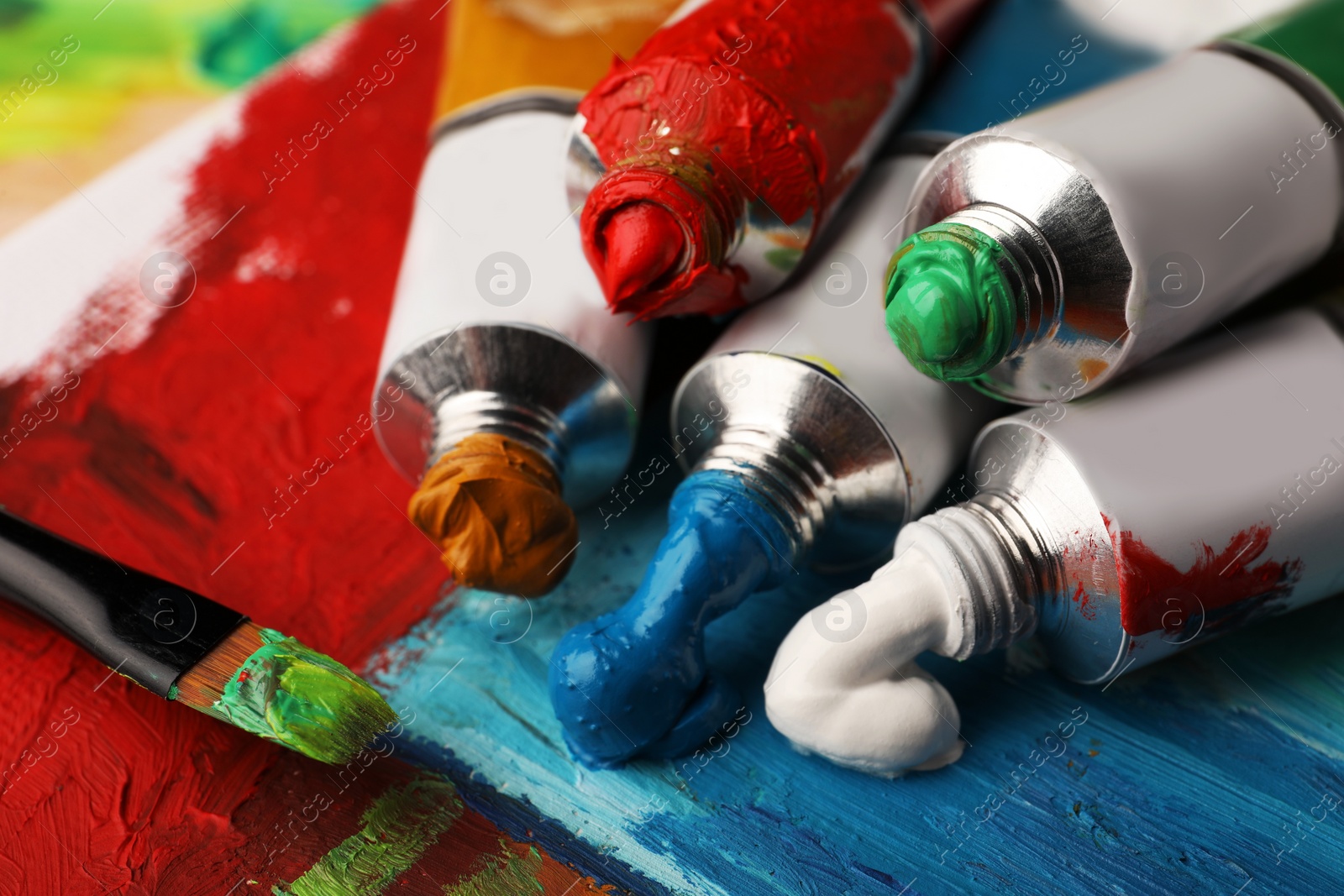 Photo of Tubes of colorful oil paints and brush on canvas with abstract painting, closeup