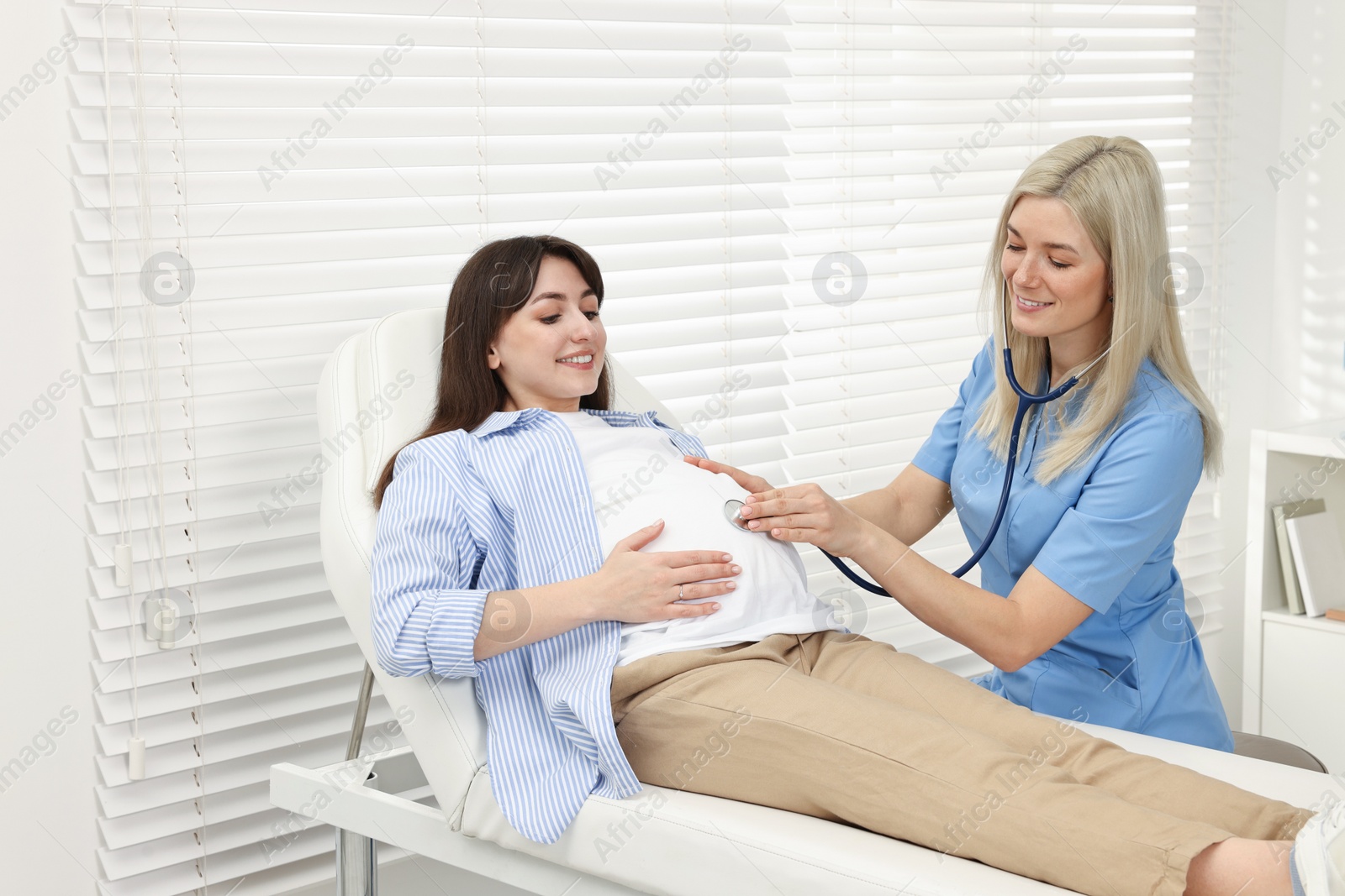 Photo of Pregnancy checkup. Smiling doctor with stethoscope listening baby's heartbeat in patient's tummy in clinic