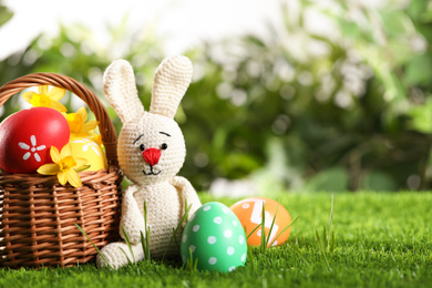 Photo of Colorful Easter eggs, toy rabbit and daffodil flowers in green grass. Space for text