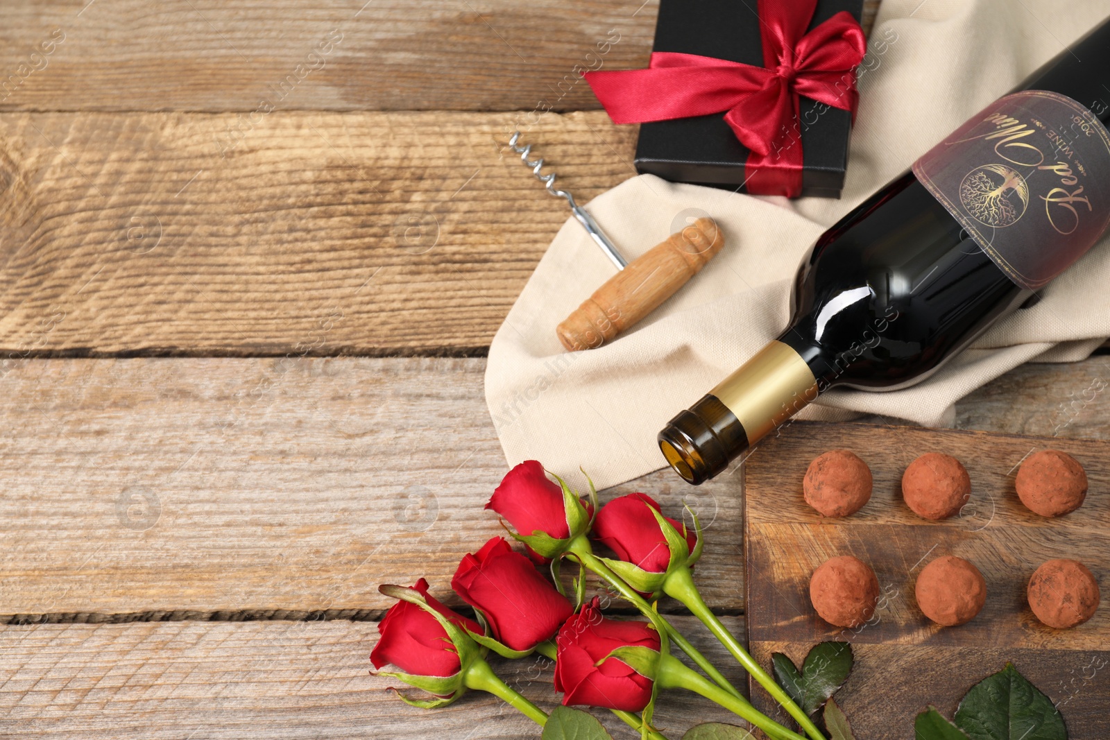 Photo of Bottle of red wine, chocolate truffles, corkscrew, gift box and roses on wooden table, flat lay. Space for text