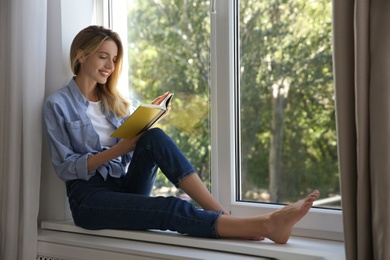Beautiful young woman reading book on window sill indoors
