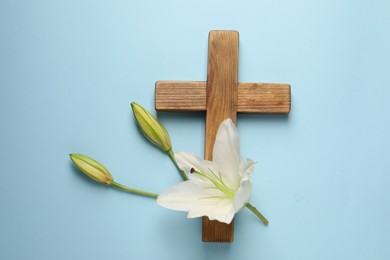 Photo of Wooden cross and lily flowers on light blue background, top view. Easter attributes