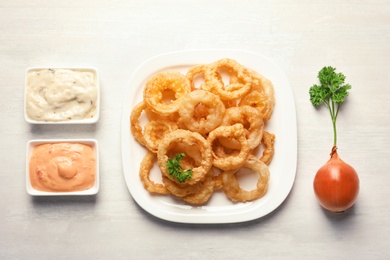 Plate with homemade crunchy fried onion rings and sauces on light background, top view