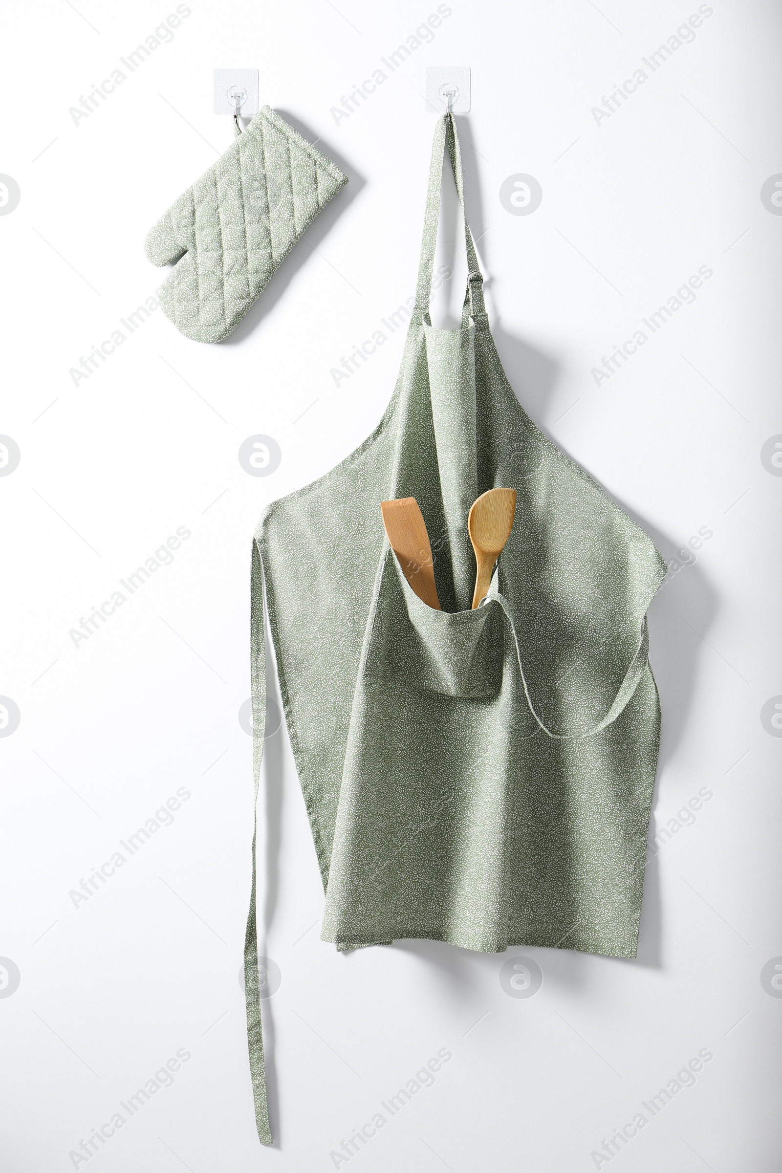 Photo of Clean apron with pattern, kitchen tools and oven glove on light wall