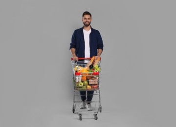 Photo of Happy man with shopping cart full of groceries on light grey background