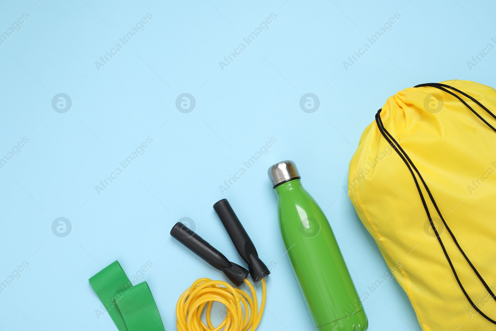 Photo of Yellow drawstring bag, thermo bottle, skipping rope and fitness elastic band on light blue background, flat lay. Space for text