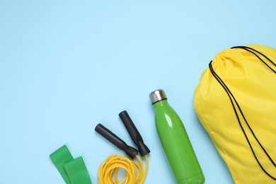 Photo of Yellow drawstring bag, thermo bottle, skipping rope and fitness elastic band on light blue background, flat lay. Space for text