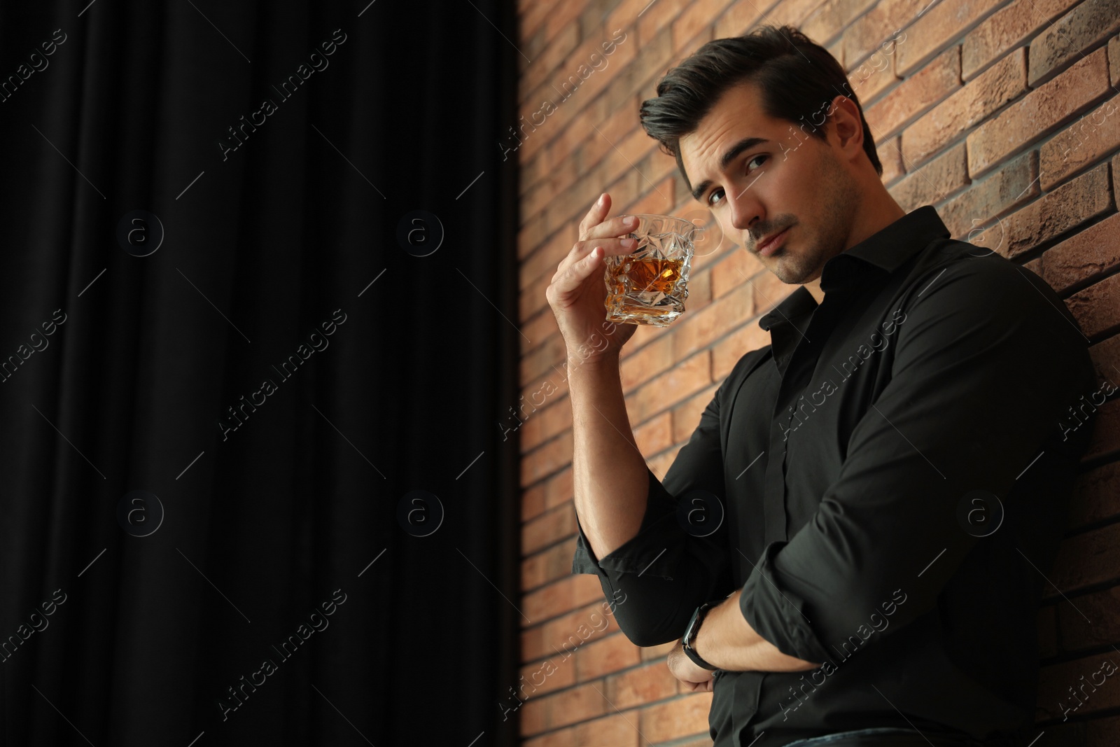 Photo of Young man with glass of whiskey near brick wall indoors. Space for text