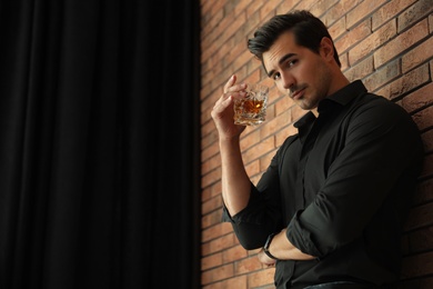 Young man with glass of whiskey near brick wall indoors. Space for text