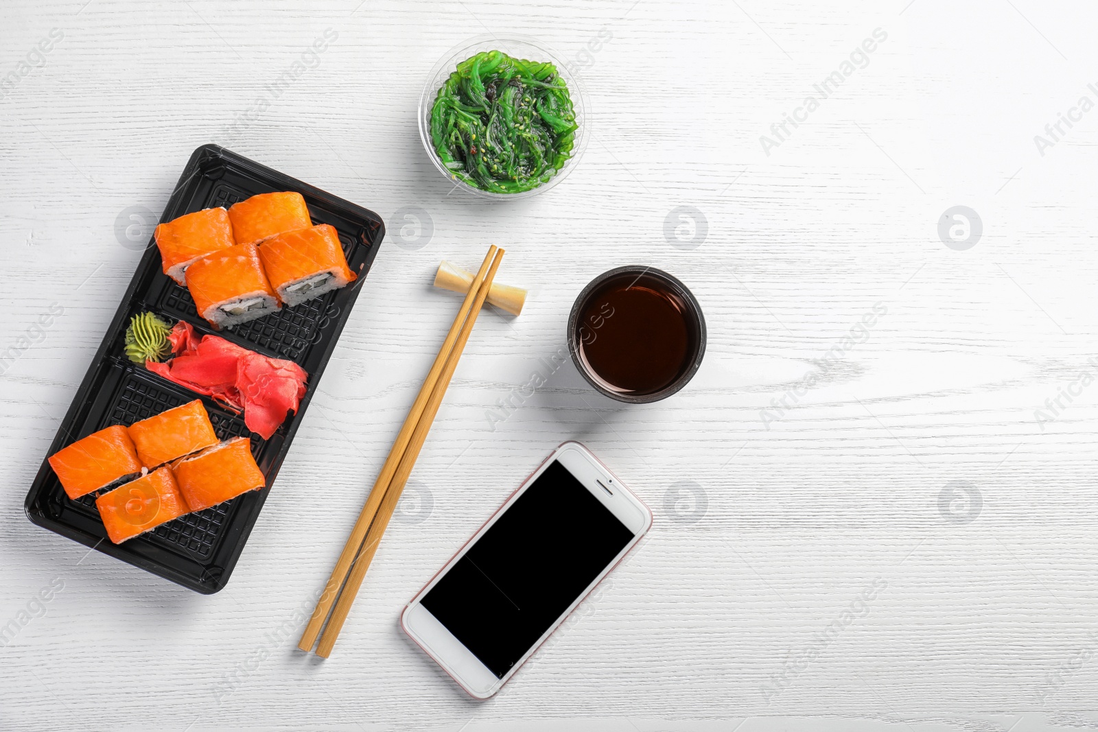 Photo of Flat lay composition with sushi rolls, smartphone and space for text on white wooden table. Food delivery