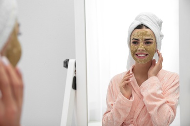 Photo of Young woman with clay mask on her face near mirror in bathroom. Skin care