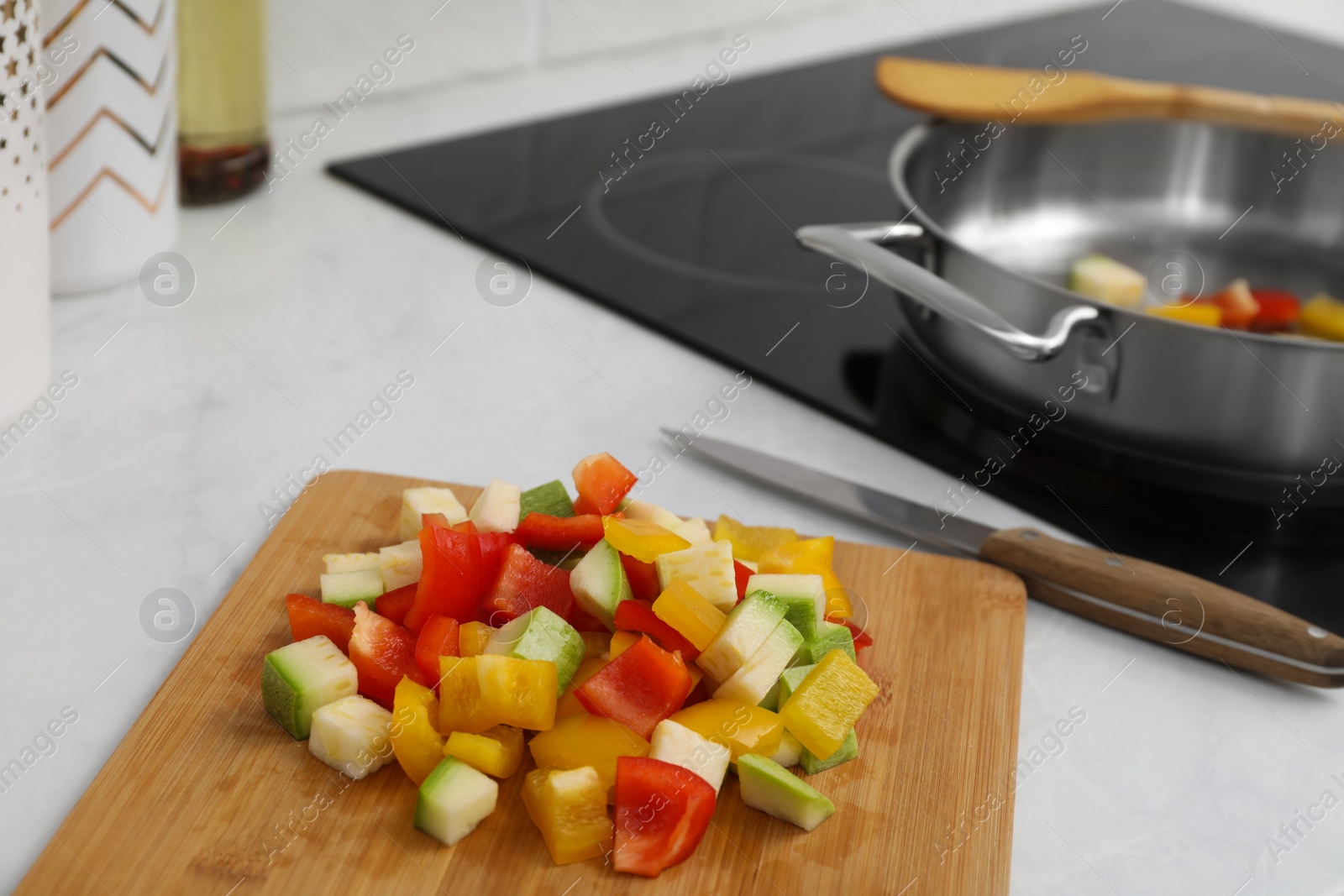 Photo of Wooden board with cut vegetables and knife near saute pan in kitchen, closeup