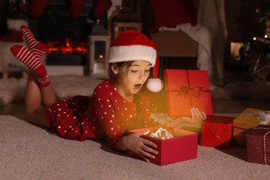 Photo of Surprised child in Santa hat opening Christmas gift on floor at home