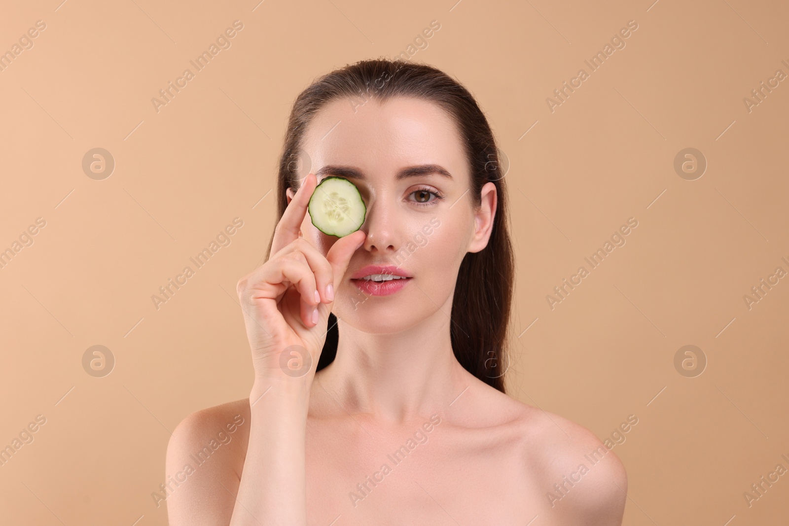 Photo of Beautiful woman covering eye with piece of cucumber on beige background