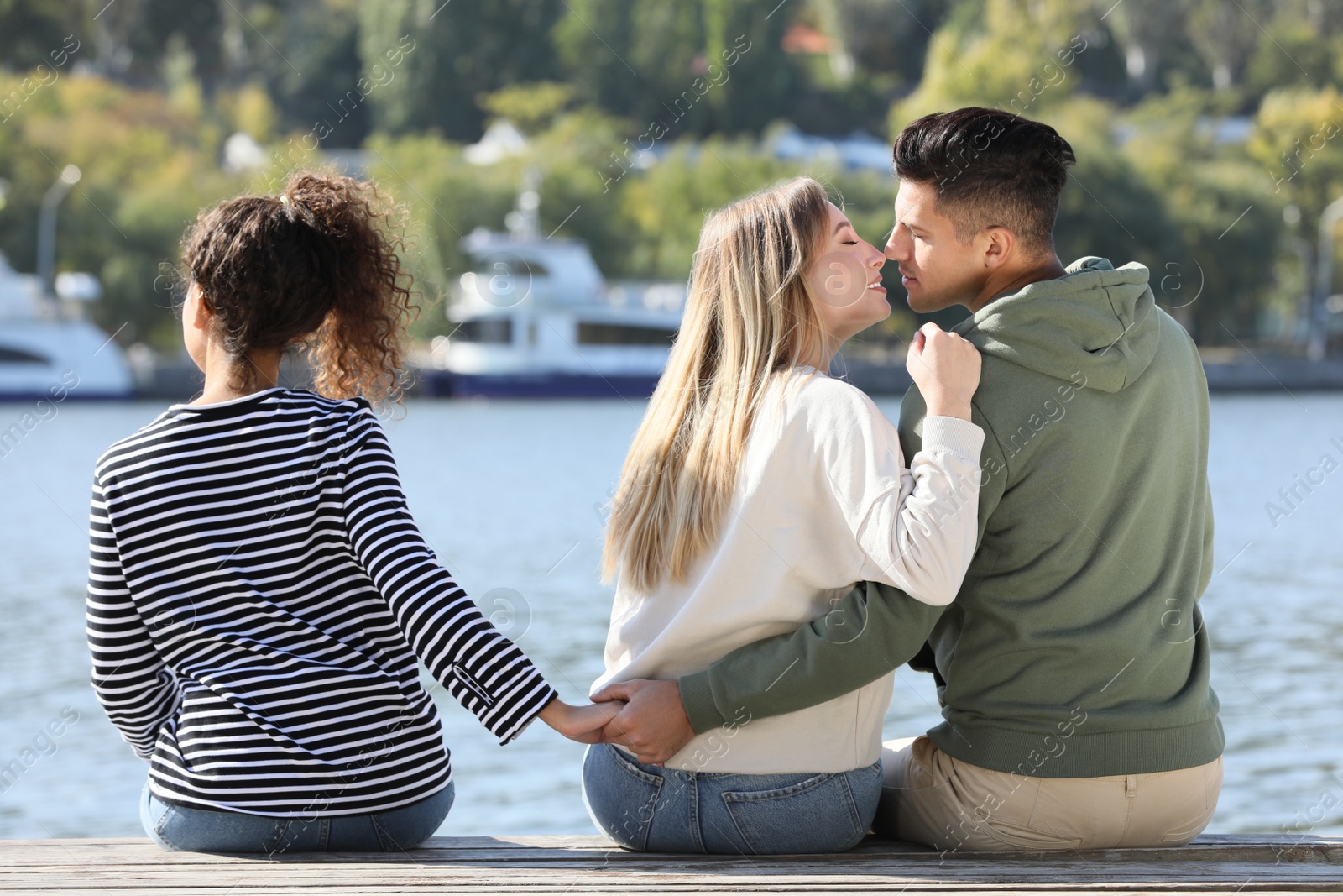 Photo of Man holding hands with another woman behind his girlfriend's back on pier near river. Love triangle