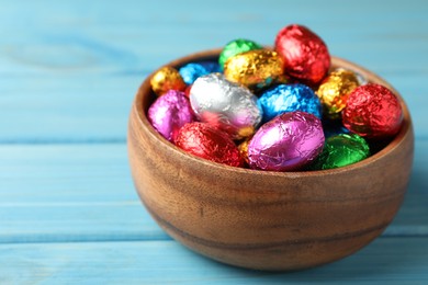 Photo of Bowl with chocolate eggs wrapped in colorful foil on light blue wooden table, closeup