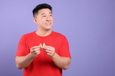 Asian man holding tasty fortune cookie with prediction on violet background. Space for text