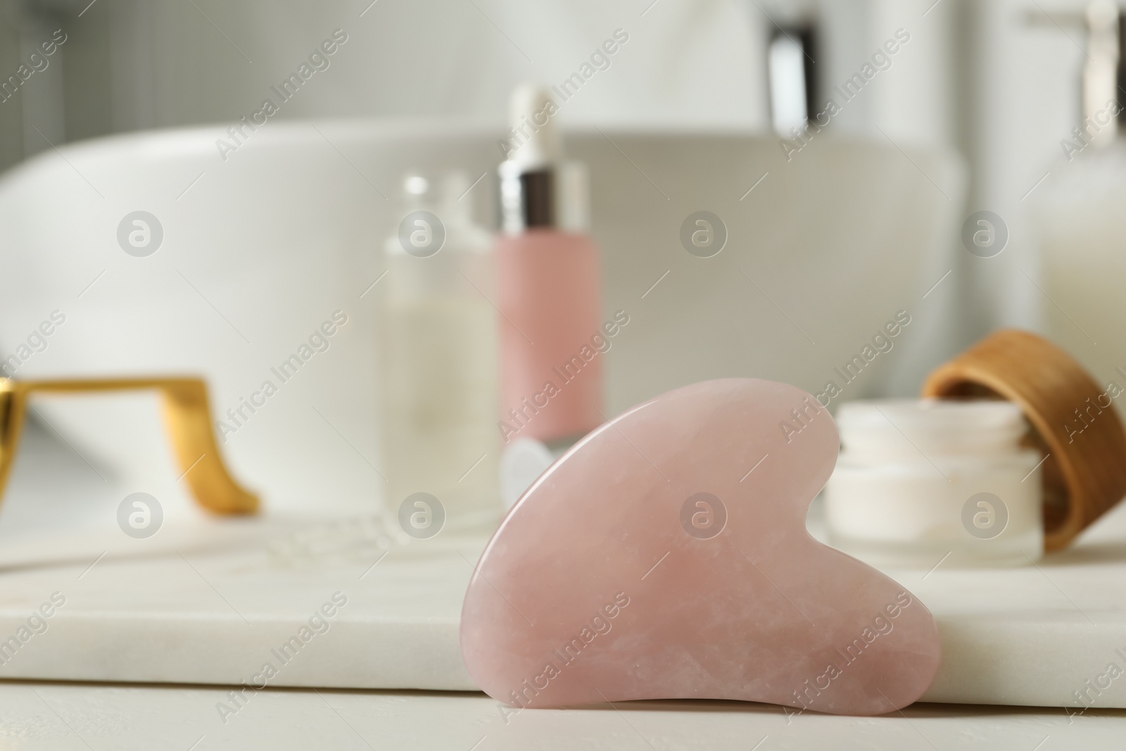 Photo of Rose quartz gua sha tool and toiletries on white countertop in bathroom, closeup. Space for text