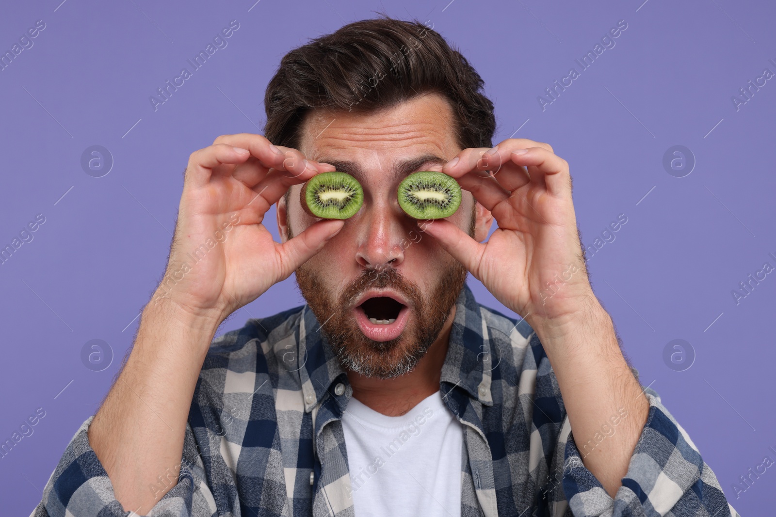 Photo of Emotional man holding halves of kiwi near his eyes on purple background