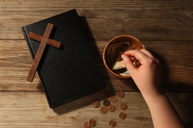 Donate and give concept. Woman putting coin into bowl with money, closeup. Bible and cross on wooden table, top view