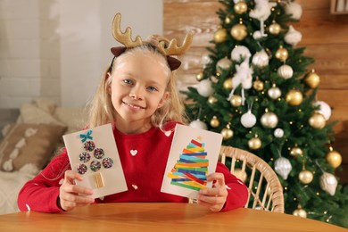 Photo of Cute little child with beautiful Christmas cards at table in decorated room