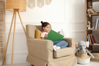 Photo of Little girl reading book in armchair at home