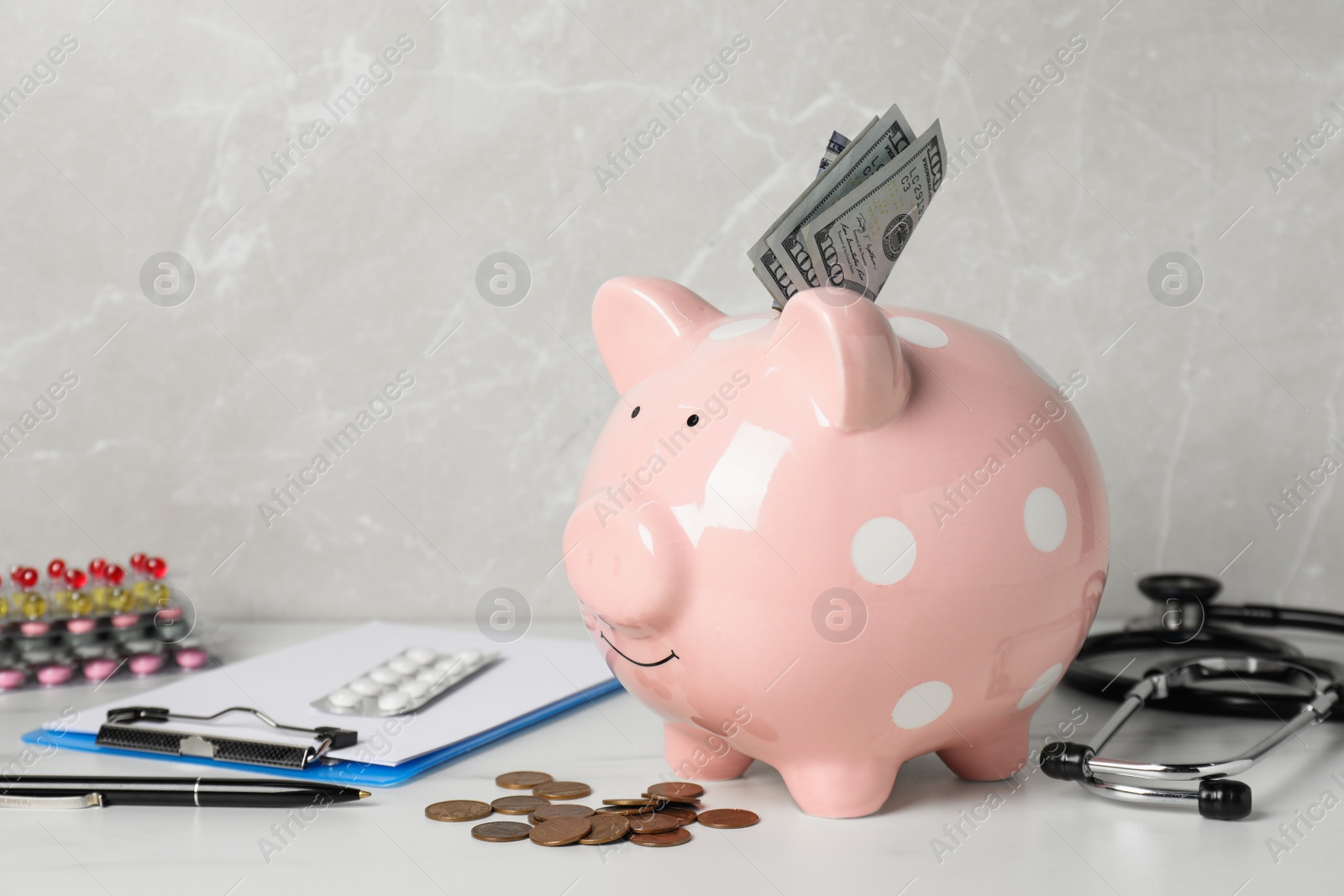 Photo of Pale pink ceramic piggy bank with money, stethoscope and pills on white table. Medical insurance