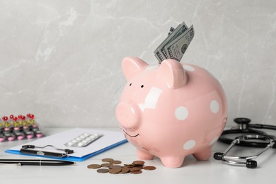 Pale pink ceramic piggy bank with money, stethoscope and pills on white table. Medical insurance