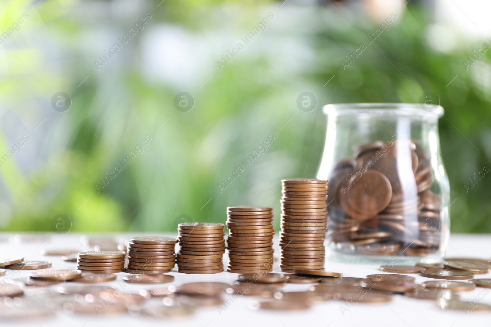 Photo of Glass jar and metal coins on white table against blurred green background. Space for text