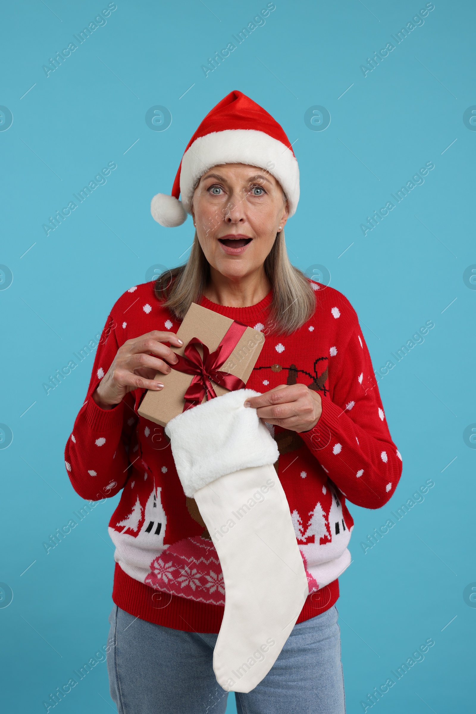 Photo of Surprised senior woman in Christmas sweater and Santa hat taking gift from stocking on light blue background