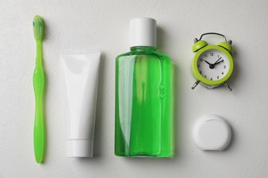 Fresh mouthwash in bottle, toothbrush, toothpaste, dental floss and alarm clock on light background, flat lay