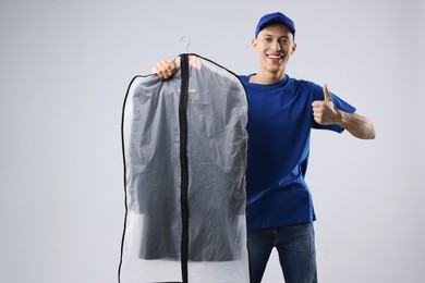 Photo of Dry-cleaning delivery. Happy courier holding garment cover with clothes and showing thumbs up on light grey background
