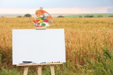 Photo of Wooden easel with blank canvas and painting equipment in field. Space for text