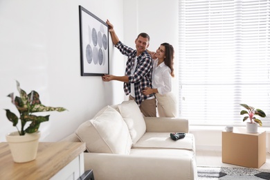 Photo of Happy couple hanging picture on white wall together. Interior design