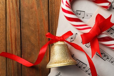 Golden shiny bell with red bow, music sheets and candy cane on wooden table, flat lay. Space for text. Christmas decoration