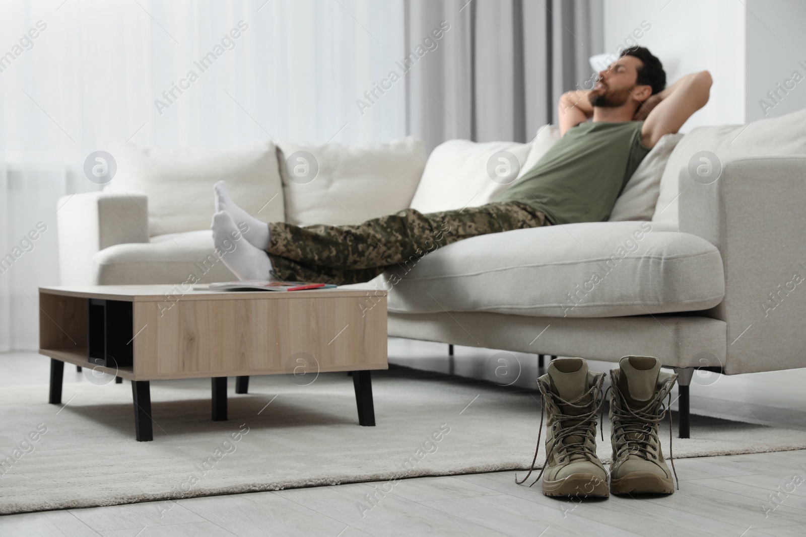 Photo of Soldier relaxing on sofa in living room, focus on pair of combat boots. Military service